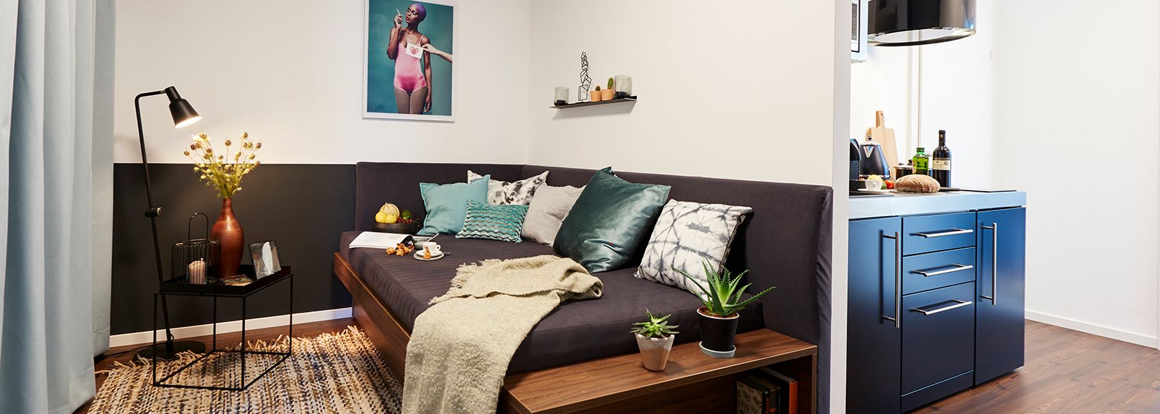 Wohnbereich eines Business-Apartments mit Sofa, Stehleuchte, Pflanzen und blauer Küchenzeile