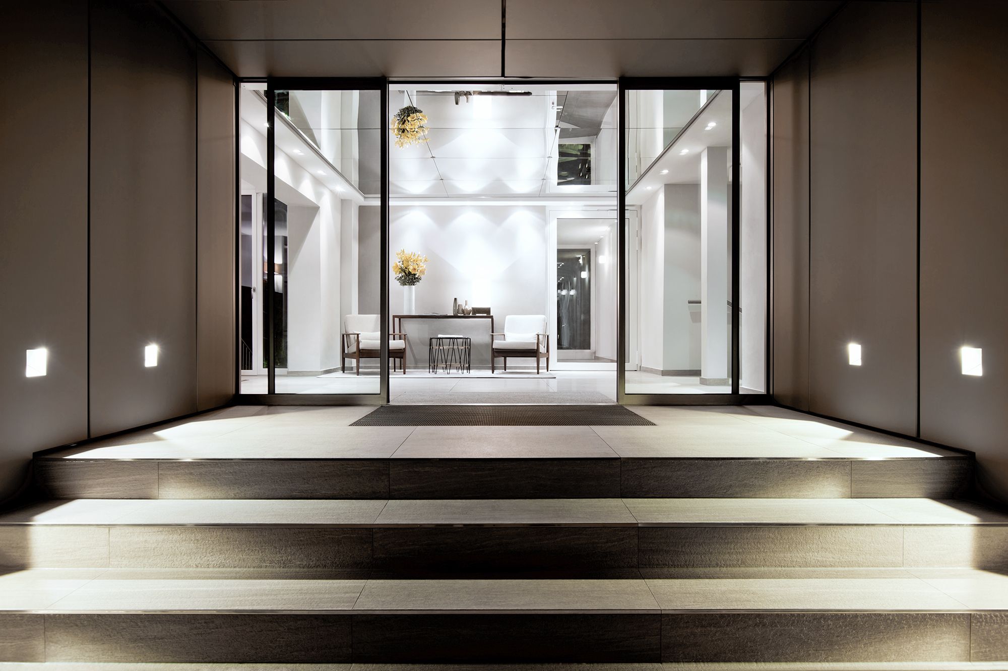 Beleuchteter Eingang mit Freitreppe, Glasfront und heller Lobby mit Sitzgruppe